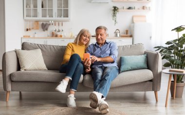Happy Mature Husband And Wife Using Smartphone With New Application, Browsing Internet Together Sitting On Sofa At Home. Senior People Texting On Phone. Mobile App, Communication And Gadgets