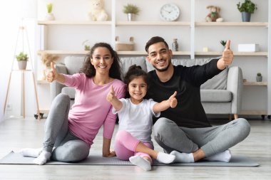 Sportif şirin bir aile, orta doğulu bir baba, anne ve küçük kızı spor kıyafetleriyle evde birlikte egzersiz yapıyor yoga minderinde oturuyor, gülümsüyor ve baş parmaklarını kaldırıyor, oturma odasının içini gösteriyor, fotokopi çekiyor.