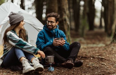 Glad young european couple tourists in jackets rest in autumn forest near tent, enjoy cold season outdoor, warming hot drink. Coffee, relax and active lifestyle, travel, adventure and camping in wood