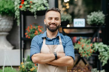 Stüdyo, kafe, dükkan sahibinin olumlu duyguları. Neşeli, kendine güvenen, önlüklü, göğsünde çapraz kolları olan sakallı yakışıklı bir adam. Çiçekçi arka planında, bitkilerle, kopyalama alanında.
