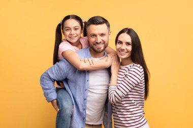 Mutlu bir aile sarı arka planda poz veriyor. Baba kızını sırtında taşırken anne ona yakın duruyor. Sıcak bir sevgi ve birliktelik ortamı yaratıyor..