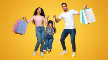 Aile alışverişi. Neşeli Arap ebeveynler ve küçük kız alışverişe çıkıyorlar ve sarı stüdyo arka planında birlikte eğleniyorlar, neşeli anne, baba ve çocuk mevsimlik satışların keyfini çıkarıyorlar.