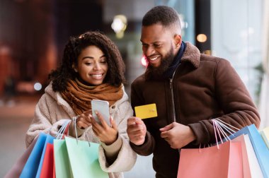 Kolay Ödeme Konsepti. Cep telefonu kullanan, kredi kartı ve alışveriş torbaları tutan ve akşam vakti dışarıda bekleyen mutlu Afro-Amerikan erkek ve kadın portresi. Perakende ve Satın Al