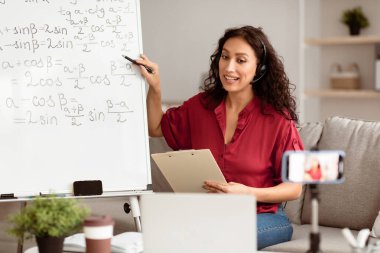 Covid-19 salgını sırasında internetten çalışma. Kulaklıklı gülümseyen matematik öğretmeni akıllı telefon ve bilgisayar kullanarak internet üzerinden ders veriyor. Vlog kaydeden öğrencilerle video görüşmesi yapan Milenyum Öğretim Üyesi