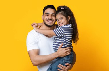 Küçük kızını elinde tutan mutlu genç bir Arap babanın portresi, Orta Doğulu sevimli bir babayla tatlı bir kız çocuğu birbirlerine bağlanırken sarı stüdyo arka planında poz veriyorlar, uzay kopyası.