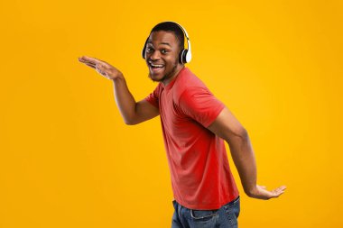 Komik siyah erkek, kablosuz kulaklıkla müzik dinliyor ve Mısır usulü dans ediyor. Sarı arka planda kameraya gülümsüyor. Stüdyo çekimi. Müzik Uygulaması Konsepti