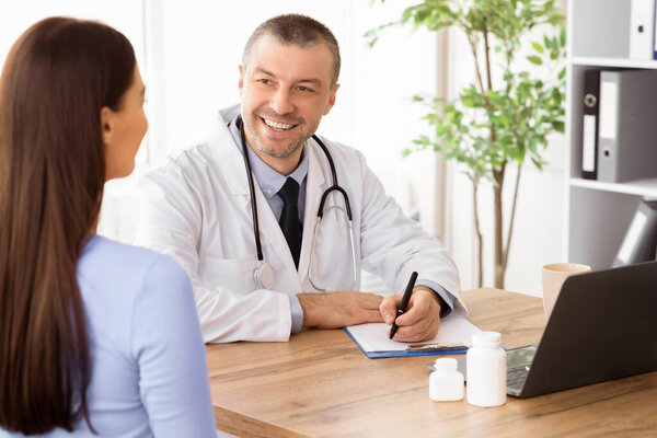 General Practitioner Appointment. Portrait of smiling doctor talking to his patient, discussing diagnosis and treatment, writing. Specialist giving consultation to woman, sitting at desk in his office