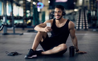 Egzersiz Uygulaması. Spor salonundaki eğitimden sonra Smartphone kullanan mutlu Arap erkek sporcu, gülümseyen Orta Doğulu adam cep telefonuyla yerde dinleniyor, internete bakıyor ya da favori müziği dinliyor, Space 'i kopyalıyor.