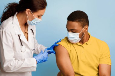 Coronavirus aşısı. Siyah Adam, Covid-19 'a karşı aşı oluyor, Doktor, Mavi Stüdyo' nun arka planına kaslar arası enjeksiyon yapan, maske takan,