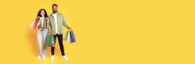 Alışverişe. Kredi kartı ve alışveriş poşetleriyle gülümseyen orta doğulu çift, stüdyoda sarı arka planda sezonluk satışlar sırasında bir sürü alışveriş satın almış. Tam boy çekim