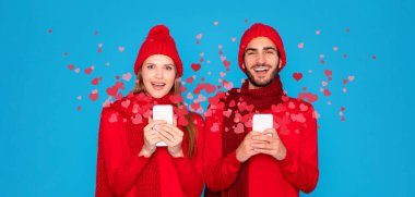 A couple in matching red outfits, wearing warm hats and scarves, smiles happily while holding smartphones. They are surrounded by floating hearts, symbolizing love and celebration on Valentines Day. clipart