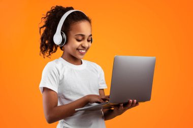 E-öğrenme kavramı. Sarı turuncu stüdyo arka planında dizüstü bilgisayarı olan, kablosuz kulaklık takan, klavyede yazan gülümseyen Afrikalı Amerikalı genç kızın portresi.