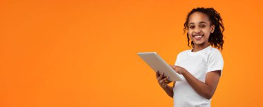 Dijital Yerliler, Çocuklar İçin Uygulama. Güzel gülümseyen Afro-Amerikan genç kız, dijital tableti turuncu stüdyo arka planında tutuyor ve kullanıyor. Kameraya bakıyor ve poz veriyor.