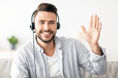 İnternetteki öğretmenlerin portresi. Kulaklık takıp web kamerasına el sallayan mutlu ve yakışıklı bir adam. Evdeki kanepede otururken selam veriyor ve gülümsüyor. Modern uzaklık öğretim ve e-öğretim kavramı