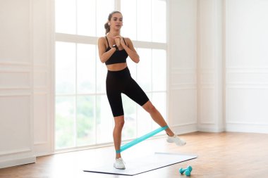 Tam ve Alt Vücut Çalışması. Elastik Lastik Bant Kaldırma ve Bacak Tekmeleme Egzersizinde Yoga Mat 'te durup Kablosuz Kulaklıklarda Müzik Dinlerken Odaklanmış Kadın Egzersizi. Vücut Ağırlığı Çalışması