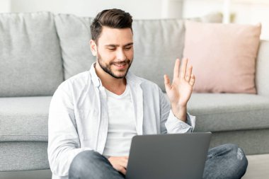 Mutlu milenyum adamı dizüstü bilgisayara el sallıyor. Oturma odasında otururken arıyor. Boş yer var. Evde bilgisayar kullanarak iletişim kuran yakışıklı bir adam.