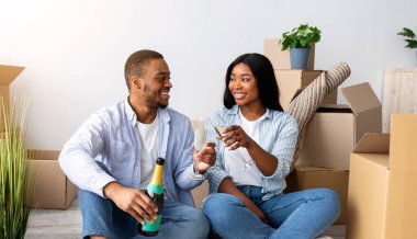 Genç Afrikalı Amerikalı çift yeni daireye taşınmayı kutluyor, karton kutuların arasında oturuyor ve şampanya içiyorlar. Ev sahipleri ve aile konutları