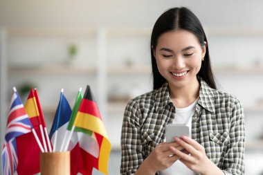Neşeli Asyalı genç kadın cep telefonu kullanıyor, İngilizce ya da diğer yabancı dil derslerine giriyor, evdeki kablosuz kulaklıklar ve uluslararası bayraklarla masada oturuyor, mobil uygulama kullanıyor, uzay boşluğunu kopyalıyor.