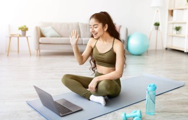 Spor giyimli genç Hintli kadın spor minderinde oturuyor, kişisel antrenörünü internette karşılıyor, bilgisayarında, içeride. Milenyum Asyalı kadın internet üzerinden iletişim kuran yoga kursuna hazır.