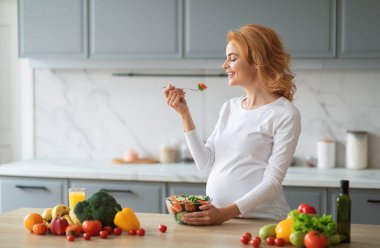 Neşeli hamile bir kadın şık bir mutfakta sağlıklı bir salatanın tadını çıkarırken gülümser. Etrafını taze sebzeler ve meyveler sarmış. Hamilelik boyunca besleyici yiyecekler üzerine odaklanmışlar..