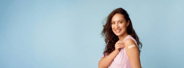 Coronavirus Aşı Reklamı. Covid-19 Aşı Pozisyonundan sonra Koluna Alçı Bandajlı Mutlu Aşılanmış Kadın Mavi Arkaplanda Gülümsüyor. Panorama, Boşluk Boşluğu