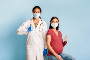 Başarılı Coronavirus aşısı. Aşılanmış Hamile Kadın ve Hemşire Covid-19 aşısı yaptırdıktan sonra başparmak hareketi yaparak mavi arka planda maske takıyor. Kapsamlı Dokunulmazlık Konsepti