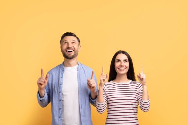 Mutlu bir çift, heyecan verici bir şekilde yukarıyı işaret ederek yan yana durur. Gülücüklerini paylaşıyorlar ve heyecanlanmış görünüyorlar, sevgilerini gösteriyorlar. Parlak sarı arkaplan ana canlı bir dokunuş katıyor..