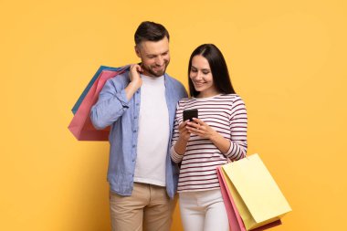 Genç bir çift birbirine yakın duruyor, akıllı telefona bakarken neşeli bir anı paylaşıyorlar. Renkli alışveriş çantaları taşıyorlar, eğlenceli günlerini sergiliyorlar..