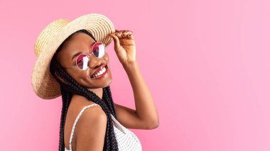 Süslü elbise ve güneş gözlüğü takan neşeli siyah bayan hasır şapkaya dokunuyor ve pembe stüdyo arka planında kameraya gülümsüyor. Pozitif Afro-Amerikalı kadın havalı kıyafetler içinde poz veriyor.