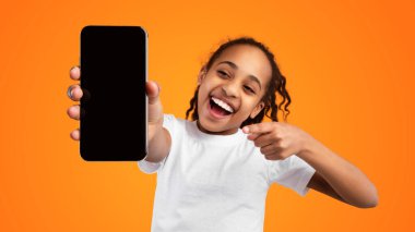 Moda Cep Telefonu. Elinde boş ekranla akıllı telefonu tutan heyecanlı genç zenci kız kameraya yakın bir cihaz gösteriyor ve boş alan, model, sarı stüdyo arka planı olan cihaza işaret ediyor.
