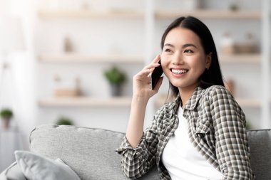 Kanepede oturan neşeli Asyalı kadın kız arkadaşı ya da sevgilisiyle akıllı telefonda konuşuyor fotokopi odasına bakıyor ve gülümsüyor. Güzel genç bir kadın ev içi telefon görüşmesi yapıyor.