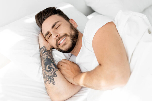 Happy morning. Satisfied guy waking up, feeling rested, happy bearded black guy lying on white linen flax bedsheets and smiling, relaxing in bedroom