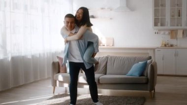 Neşeli bir çift, eğlenceli bir etkileşime girer ve bir eş içeride sırtına biner. Güneşli oturma odasında modern bir dekor ve rahat bir atmosfer var..