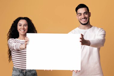 Reklam afişi. Beyaz boş levha sergileyen neşeli genç Arap çift, Orta Doğulu Mutlu Adam ve Kadın, Sarı arka planda dururken Boş Poster Tutuyor, Boşluğu Kopyala