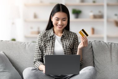 Neşeli genç Asyalı kadın oturma odasında kanepede oturuyor ve dizüstü bilgisayar ve kredi kartı kullanıyor, yeni web mağazasında online alışveriş veya bankacılık, ev içi, fotokopi alanı, satış yakalama
