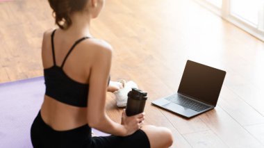 Yoga minderinde oturan, su içen, boş dizüstü bilgisayara bakan, antrenörle görüntülü konuşma yapan ya da spor salonuna katılan tanınmayan bir kadının omuz manzarası.