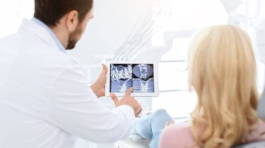 Erkek dişçi kadın hasta tedavi hedefini gösteriyor, dijital tablet kullanıyor, dişçi kliniği içi. Sakallı adam stomatolog, dişçi koltuğundaki kadınla konuşuyor. Cihazı işaret ediyor, arkadan bakıyor.