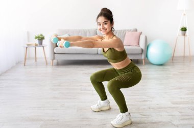 Güçlü genç Hintli kadın dambıllarla çalışıyor, evde çömelmiş, boş bir alanda. Sağlığına dikkat eden, ev içi eğitim alan, aktif bir yaşam tarzı olan sağlıklı Doğulu kadın.