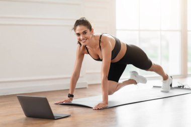 Gülümseyen Sporcu Kadın Laptop 'un önünde antrenman yapıyor, güzel bir kadın vücut dağları tırmanışı yapıyor, kameraya bakıyor ve eğitmenle çevrimiçi video görüşmesi yapıyor.