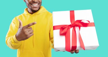 Hediye teslimatı. Tanımlanamayan Afro-Amerikalı adam elinde kırmızı fiyonklu hediye kutusuyla mavi arka planı işaret ediyor. Tatil kutlamaları, hediyeler ve hediyeler sizin için. Kırpılmış, Panorama