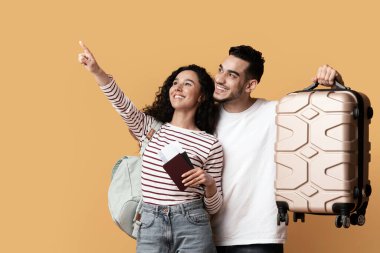 Seyahat Reklamları. Çantalı ve pasaportlu neşeli bir Arap çift sarı arka planda fotokopi alanını işaret ediyor mutlu Orta Doğulu gezginler reklam ve panorama için bedava yer gösteriyorlar.