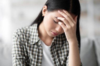 Genç Asyalı kadın baş ağrısı çekiyor, alnına dokunuyor, aşağı bakıyor, ev içi, yakın çekim, kopyalama alanı. Esmer kadın üzgün ya da migrenden muzdarip, oturma odasında kanepede oturuyor.