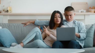 Genç bir çift gri bir kanepede rahatça oturup online alışveriş yapıyor. Kadın kredi kartı tutarken, adam dizüstü bilgisayara odaklanıyor, ikisi de rahatlamış görünüyor..