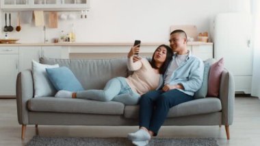 Genç bir çift rahat bir kanepede oturup selfie çekerken gülümserler. Oturma odasında açık renkler ve modern bir dekor var, rahat bir atmosfer yaratıyor..