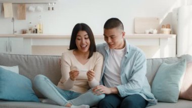 Bir çift rahat oturma odalarındaki kanepede oturup hamilelik testine bakarken heyecanlı bir anı paylaşıyorlar. Yüz ifadeleri gelecekle ilgili sevinç ve beklenti iletir..