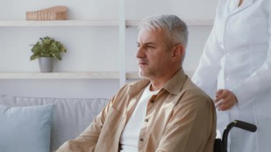 Tekerlekli sandalyedeki orta yaşlı beyaz bir adam evde rahat bir şekilde oturur. Bakıcı nazikçe elini omzuna koyar, rahat bir ev ortamında güven ve destek sağlar..