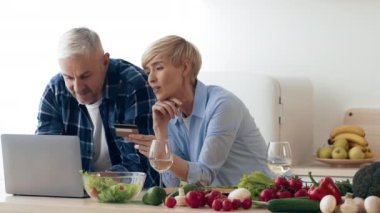 Bir çift, yemek tariflerini bulmak için dizüstü bilgisayar kullanarak mutfakta yemek hazırlıyor. Taze sebzeler ve meyveler masanın etrafını sararak sıcak ve davetkar bir atmosfer yaratıyor..