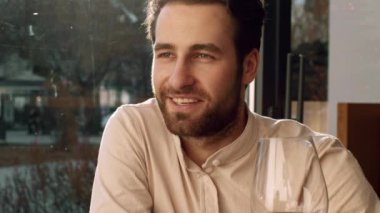 Genç bir adam rahat bir şekilde bir restoran masasında oturur, gülümser ve yemeğinin tadını çıkarırken doğal ışık pencereden içeri süzülür, öğleden sonranın rahat atmosferini ön plana çıkarır..