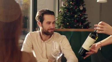 Bir adam rahat bir restoranda masada oturur ve bir garsonun şarap sunuşunu seyreder. Noel süslemeleri arka planı süsleyerek, akşamları şenlikli bir ortam yaratıyor..