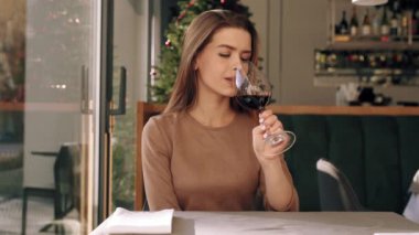 Bir kadın restoranda bir masada oturmuş düşünceli bir ifadeyle bir bardak kırmızı şarabın tadını çıkarıyor. Sıcak ortam onun rahatlamış ruh halini tamamlıyor..